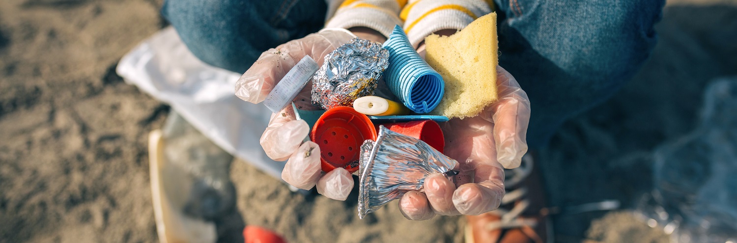 Zum Thema Abfallvermeidung kniet eine Person im Sand und hält bunten Plastikmüll in den Händen, der am Strand gefunden wurde.