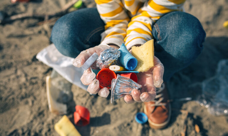 Eine Person kniet im Sand und hält bunten Plastikmüll in den Händen, der am Strand gefunden wurde.