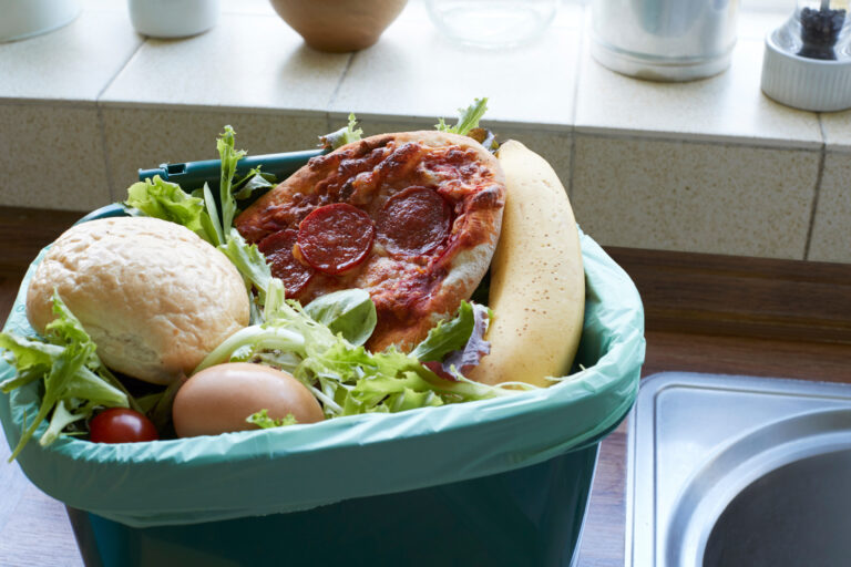 Eine kleine offene Biotonne für die Küche, gefüllt mit Abfällen wie Bananenschalen, Pizzaresten, Brotkrumen und Salatblättern.