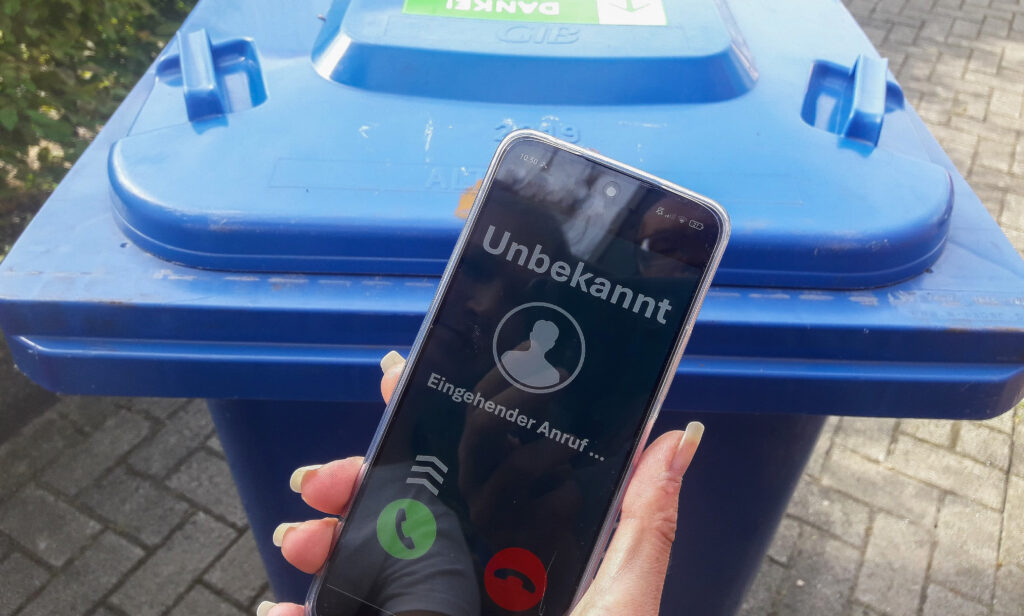 Hand hält Handy mit Unbekannten Rufeingang. Im Hintergudn ist eine Papiertonne.